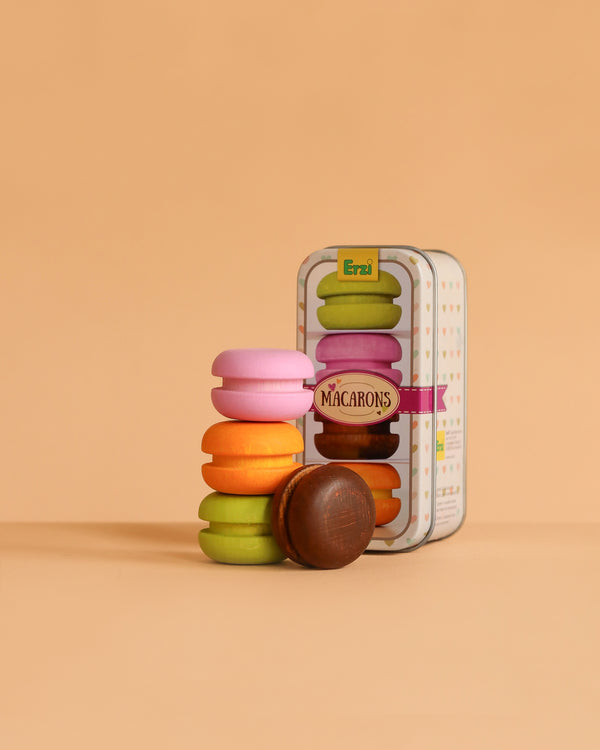 The Erzi Macarons In A Tin Pretend Food features colorful wooden macarons in pink, purple, orange, yellow, green, and brown beside a decorative "MACARONS" tin—perfect for kitchen or store play on a peach background.