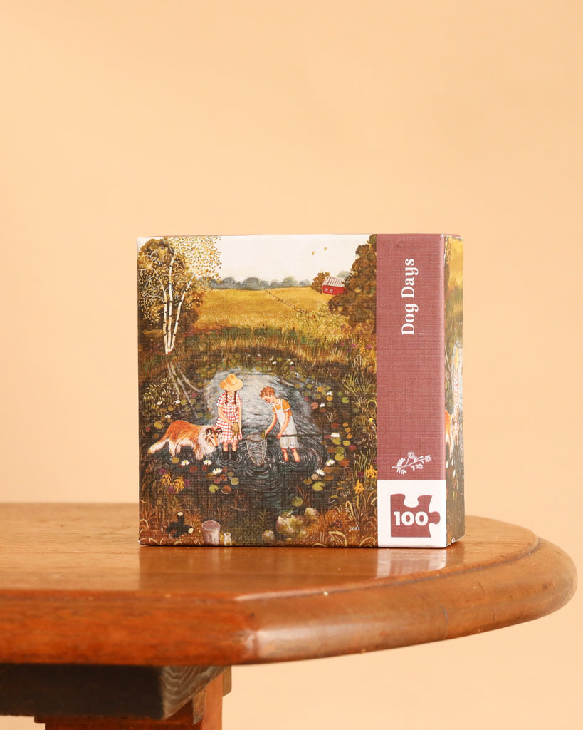 A square Dog Days Mini - 100 Piece Mini Puzzle box sits on a wooden table, featuring an illustration of two children and a dog at a pond, with the title and piece count printed on the side.