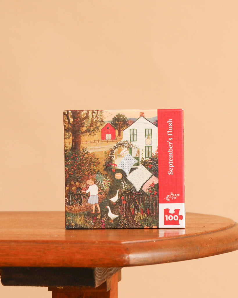 A square September’s Flush Mini - 100 Piece Mini Puzzle box featuring a folk-art illustration of a person gardening near a house, with a red side panel and label detailing the 100-piece count for this countryside jigsaw puzzle.