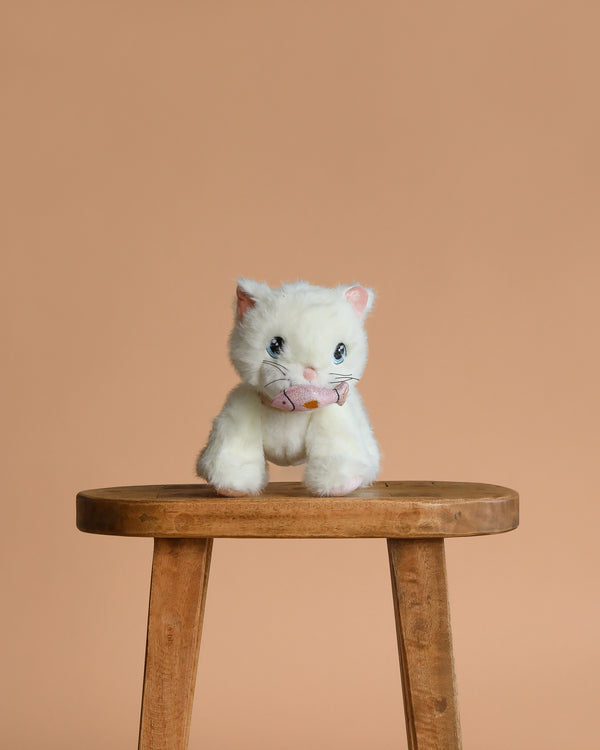 The Olli Ella Dinkum Cat Lux - White, a small posable plush kitty with blue eyes, sits on a wooden stool against a plain light brown background.
