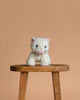 The Olli Ella Dinkum Cat Lux - White, a small posable plush kitty with blue eyes, sits on a wooden stool against a plain light brown background.