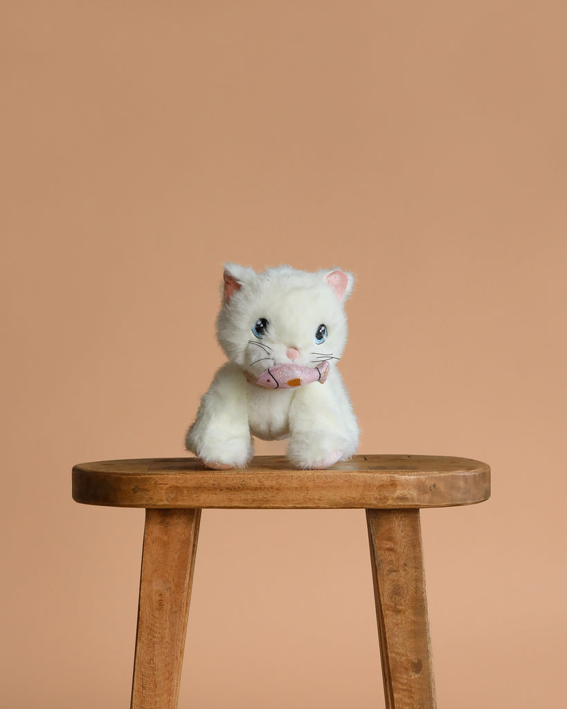 The Olli Ella Dinkum Cat Lux - White, a small posable plush kitty with blue eyes, sits on a wooden stool against a plain light brown background.