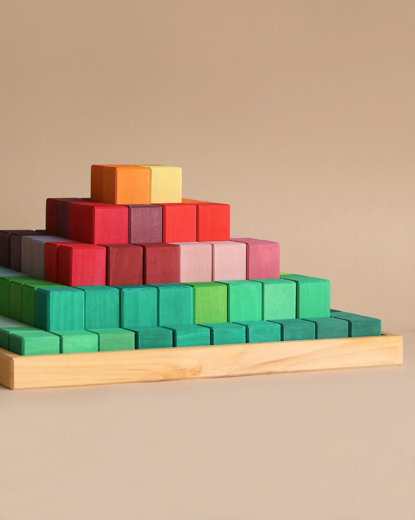 A Large Stepped Pyramid Block Set built from colorful, non-toxic wooden blocks arranged on a wooden base. The blocks are organized by color with green at the bottom, followed by blue, red, purple, orange, and a small yellow block at the top. Perfect for developing fine motor skills against a solid beige background.