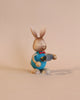 A Collectible Dregeno Easter Figure - Rabbit With Egg Carton, handcrafted in Germany, wearing a blue top and brown pants, depicted in a walking pose with one arm stretched forward and the other back, on a plain beige background.