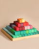 A Large Stepped Pyramid Block Set made of non-toxic wood blocks of various sizes and colors is arranged on a light wooden board. The layers transition from green at the base to blues, purples, reds, and oranges at the top. The background is a plain beige color—perfect for enhancing fine motor skills.