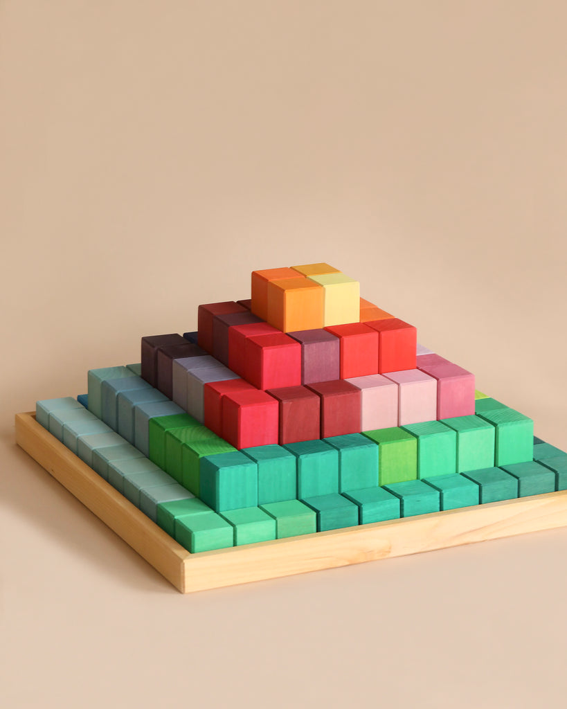 A Large Stepped Pyramid Block Set made of non-toxic wood blocks of various sizes and colors is arranged on a light wooden board. The layers transition from green at the base to blues, purples, reds, and oranges at the top. The background is a plain beige color—perfect for enhancing fine motor skills.