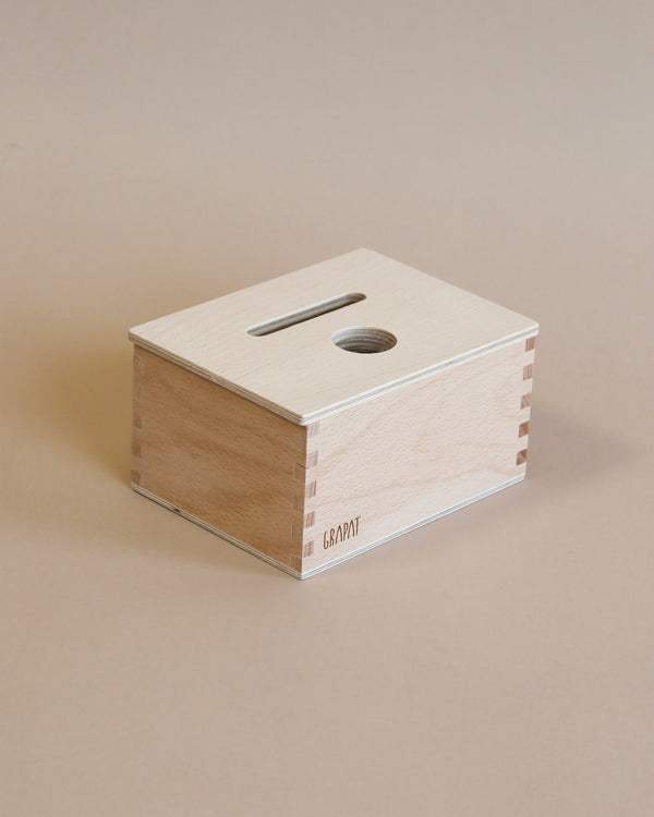 A simple, rectangular wooden box with a slot on the top, labeled "Grapat Permanence Box" on its side, resting on a beige surface. This box is crafted from wood sourced from sustainable forests.