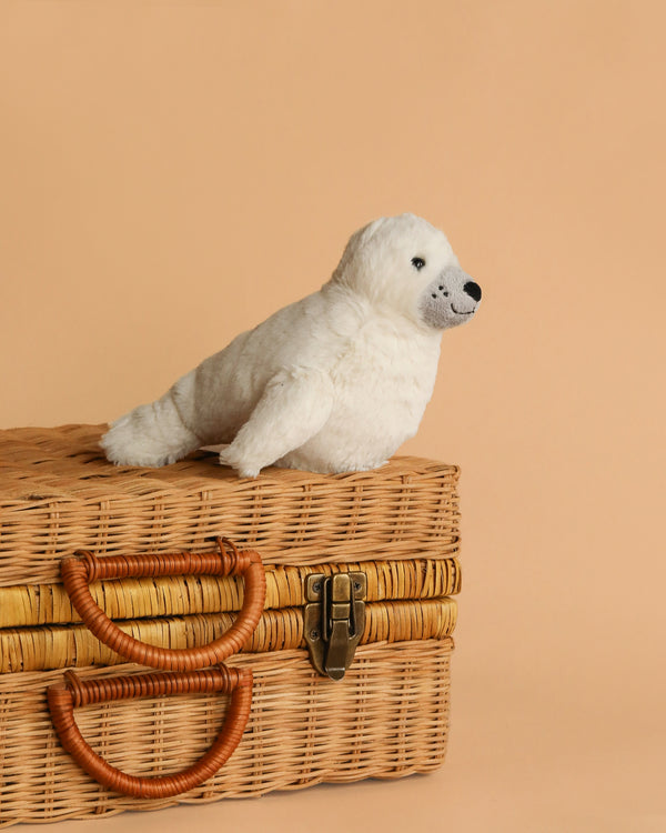 A Steiff Aro Baby Seal Pup plush sits atop a closed wicker picnic basket with leather handles, set against a plain beige background.