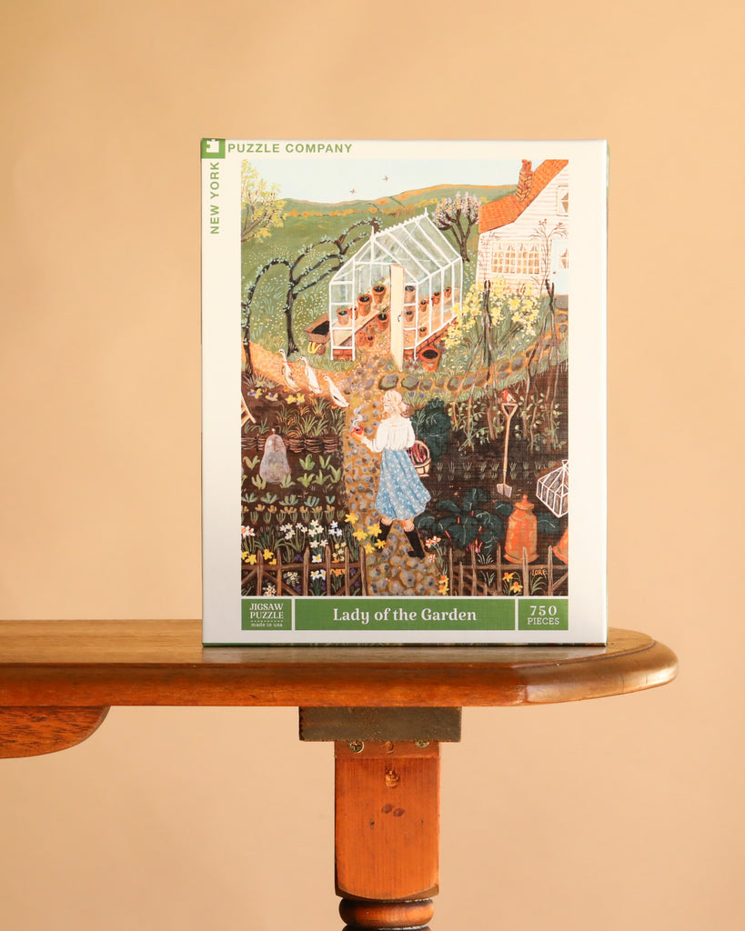 Lady of the Garden - 750 Piece Spring Jigsaw Puzzle box featuring an illustration of a woman in a blue dress walking through a garden with a greenhouse, vegetables, and flowers.