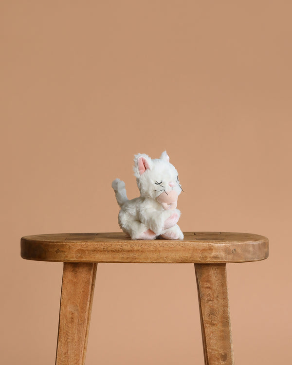The Olli Ella Binky Kitten Kiki - White, a small white and pink plush toy, sits on a wooden stool against a plain light brown background, creating an adorable display for this cuddly kitten.