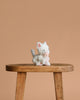 The Olli Ella Binky Kitten Kiki - White, a small white and pink plush toy, sits on a wooden stool against a plain light brown background, creating an adorable display for this cuddly kitten.