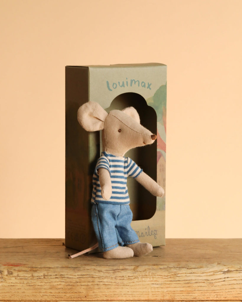 A Maileg LouisMax Mouse - Big Brother in a striped shirt and blue pants sits before a decoration box, perched on a wooden surface against a beige background, inviting imaginative journeys.