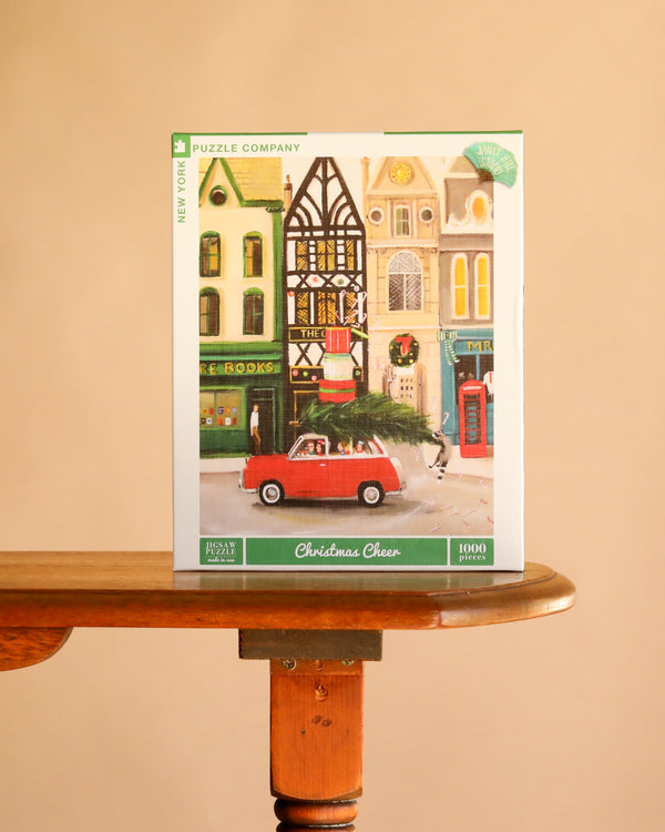 Christmas Cheer - 1000 Piece Jigsaw Puzzle box featuring a Janet Hill illustration of a red car carrying a Christmas tree through a city street lined with shops.