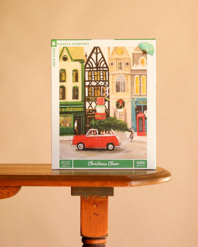 Christmas Cheer - 1000 Piece Jigsaw Puzzle box featuring a Janet Hill illustration of a red car carrying a Christmas tree through a city street lined with shops.