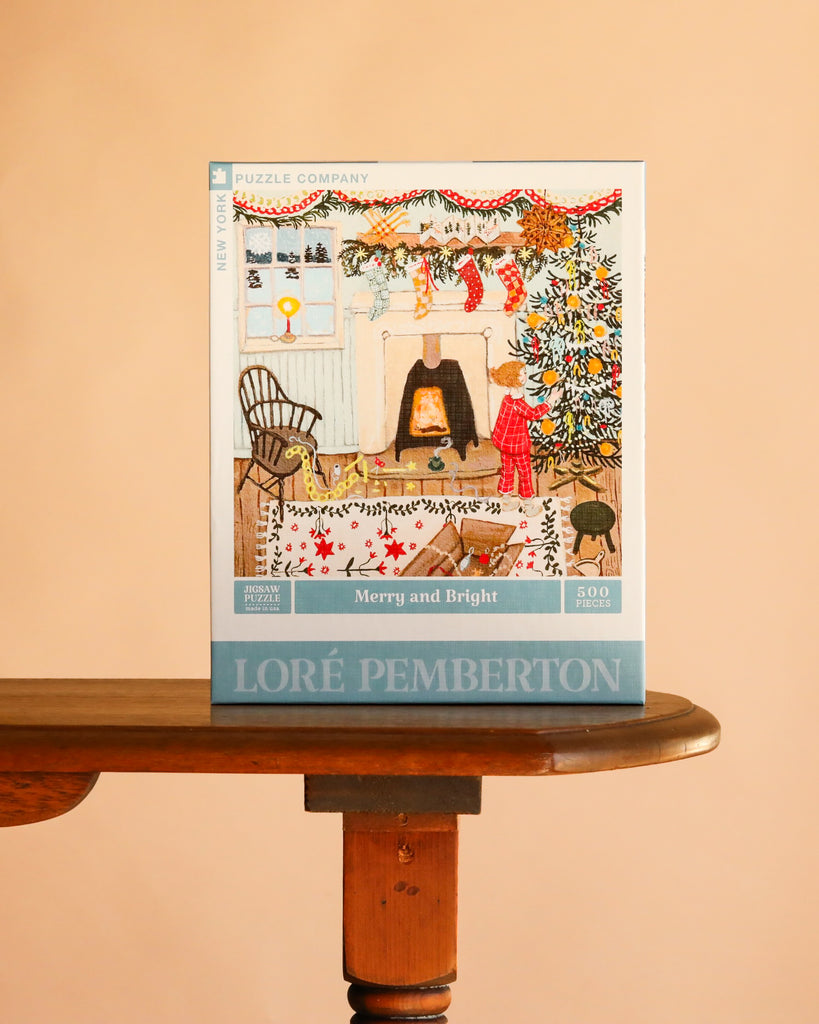 A Merry and Bright - 500 Piece Jigsaw Puzzle box showing an illustration of a child decorating a tree by a fireplace, resting on a wooden table.