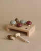 A Wooden Mushroom Hammer Board featuring mushroom-shaped pegs in various muted colors inserted into a wooden board. Two wooden mushrooms and a small mallet with a string attached are placed beside the base on a beige background.