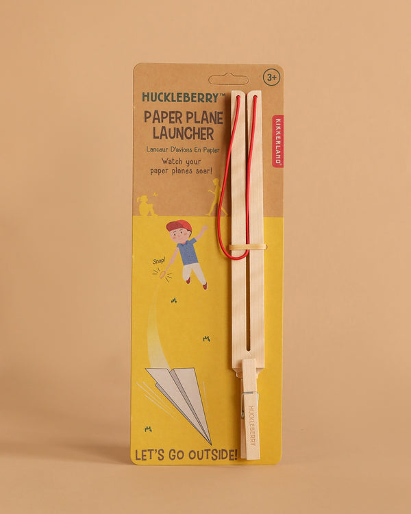 The Huckleberry Paper Plane Launcher is a birch wood set with a wooden stick and red elastic bands, designed for ages 3+, packaged with an image of a child launching a paper airplane and the phrase "LET'S GO OUTSIDE!.