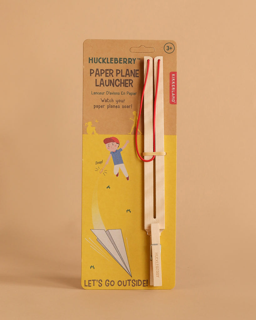 The Huckleberry Paper Plane Launcher is a birch wood set with a wooden stick and red elastic bands, designed for ages 3+, packaged with an image of a child launching a paper airplane and the phrase "LET'S GO OUTSIDE!.