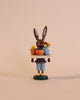 A Collectible Dregeno Easter Figure - Bunny Florist standing upright, dressed in blue overalls, handcrafted in Germany, holding a basket of colorful flowers. The bunny has a dark brown color and stands on a.