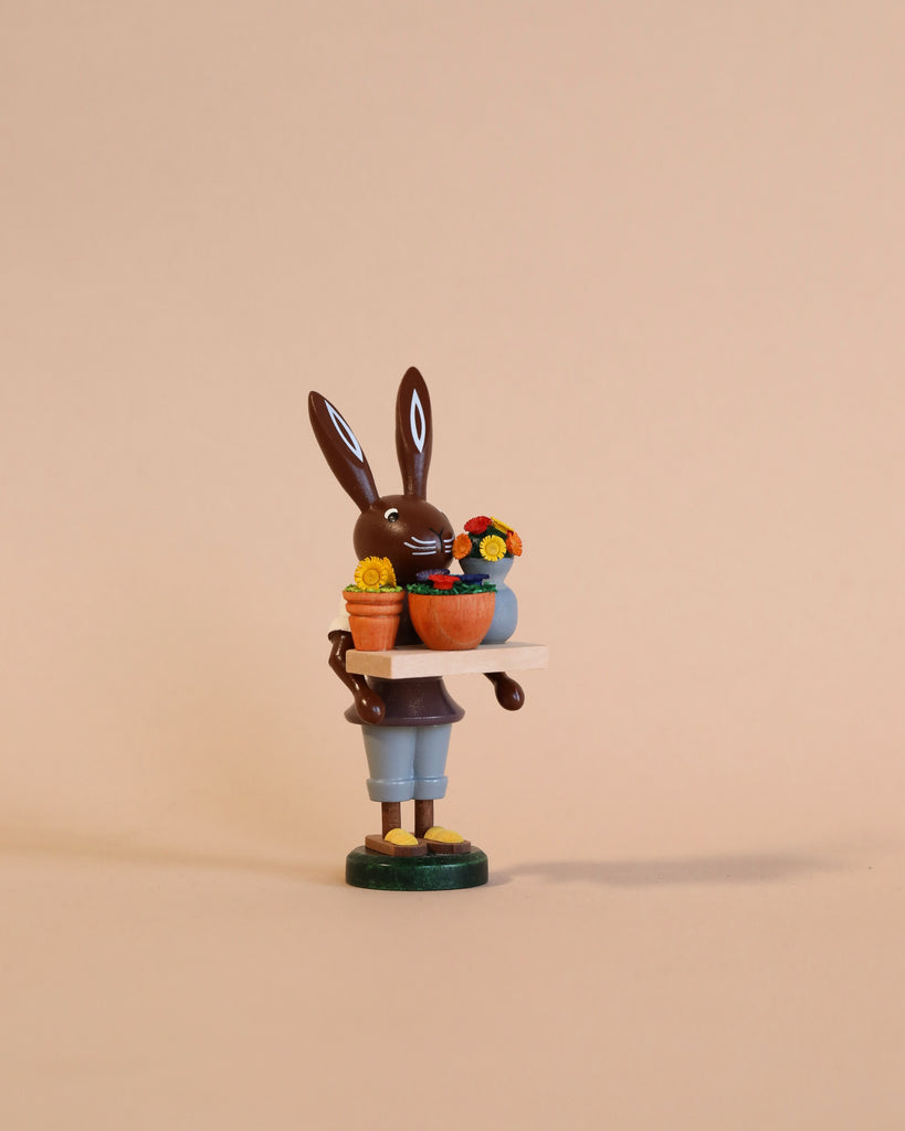 A whimsical Collectible Dregeno Easter Figure - Bunny Florist handcrafted in Germany of a chocolate bunny holding a basket of colorful flowers, posed against a soft beige background. The bunny is standing on a green circular base.