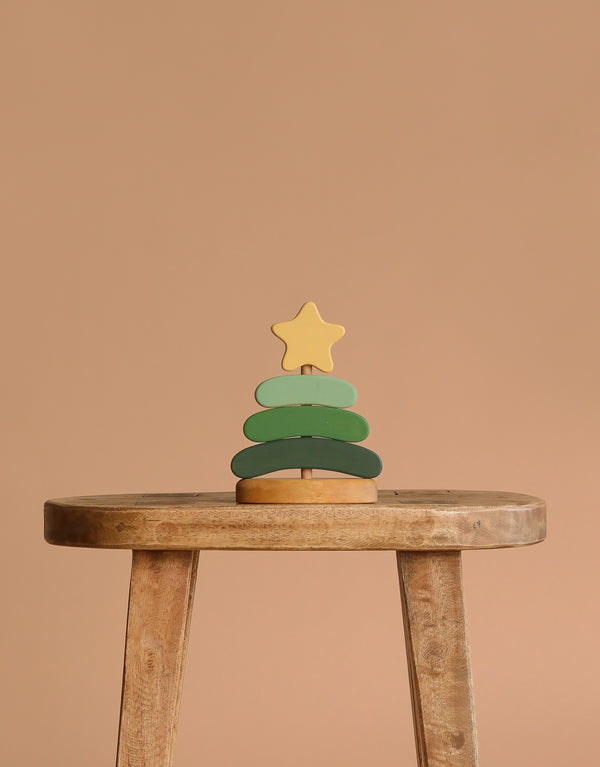 The Handmade Wooden Stacking Toy Green Christmas Tree, featuring green layers and a yellow star topper, is displayed on a rustic wooden stool against a plain beige background.
