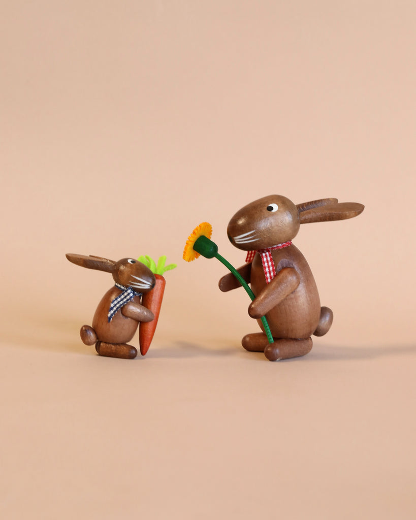 Two Collectible Dregeno Easter Figures - Bunnies With Mayflower and Carrot (Set 2), handcrafted in Germany, dressed in checkered scarves; one larger rabbit offers a small carrot to a smaller rabbit against a plain, light brown background.