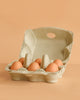 The Erzi Half A Dozen Wooden Eggs are displayed in an open cardboard carton on a light brown surface against a peach-colored background—perfect for sparking imaginative play in any pretend kitchen toy setup.