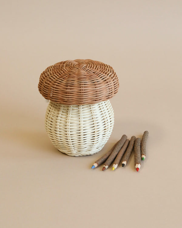 A small, whimsical Olli Ella Mushroom Basket, paired with five colorful, hand-carved wooden pencils on a neutral background. The basket has a white base and a brown top.