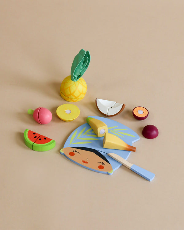 Colorful wooden play food items arranged on a beige background, including a smiling pie, a pineapple, slices of watermelon, apple, and other fruits with a tiny knife and velcro for easy cutting from the Tropical Fruit Chopping Board.