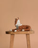 The Maileg Cosy Bambi Deer - Mint, with brown and white spotted fabric and a blue ribbon collar, rests on a wooden stool—an adorable decoration or playmate for Maileg fans against a plain beige background.