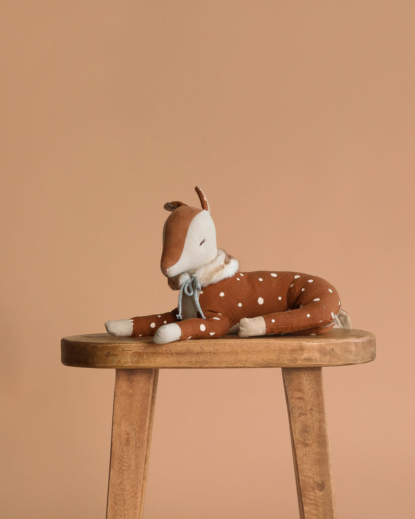 The Maileg Cosy Bambi Deer - Mint, with brown and white spotted fabric and a blue ribbon collar, rests on a wooden stool—an adorable decoration or playmate for Maileg fans against a plain beige background.