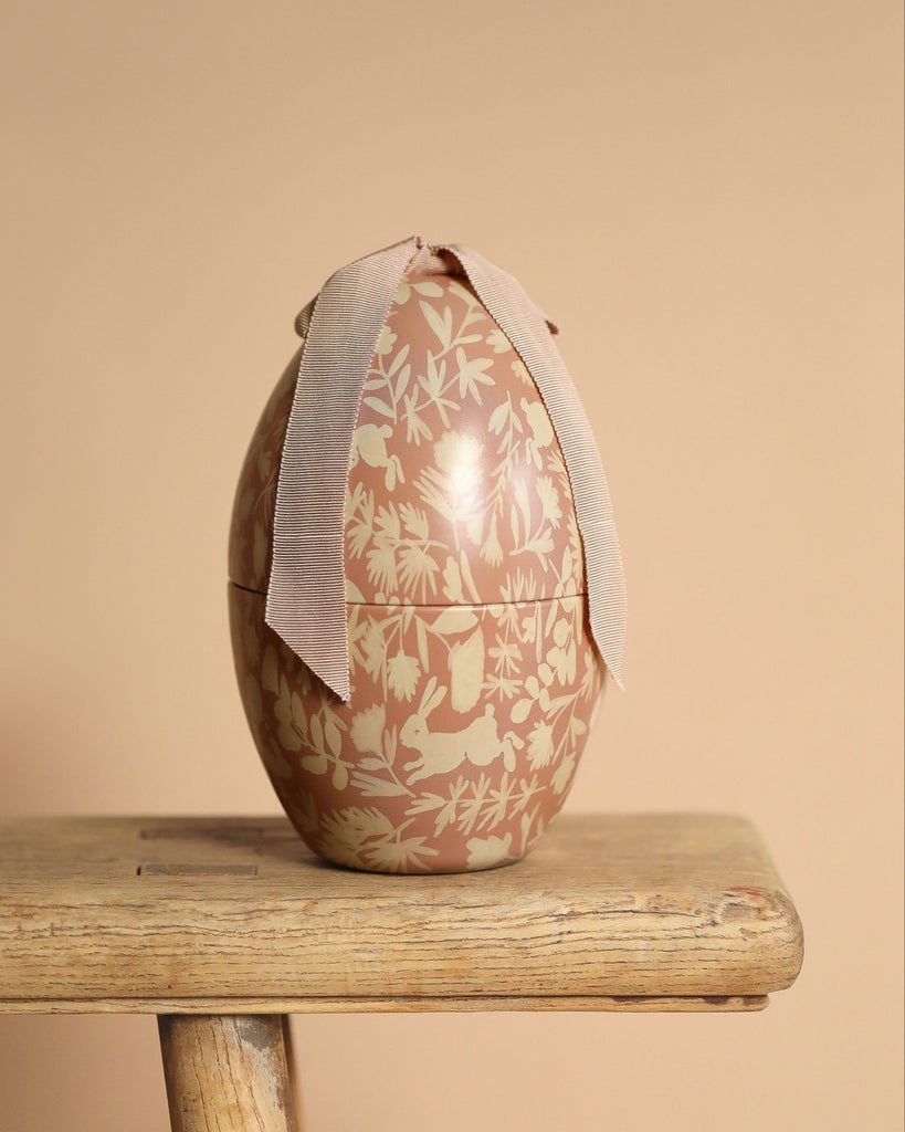 A decorative Maileg Easter Egg - Powder with a floral and leaf pattern in beige and cream tones sits elegantly on a wooden bench. Adorned with a light beige ribbon at the top, its exclusive look stands out against the soft, neutral beige background.