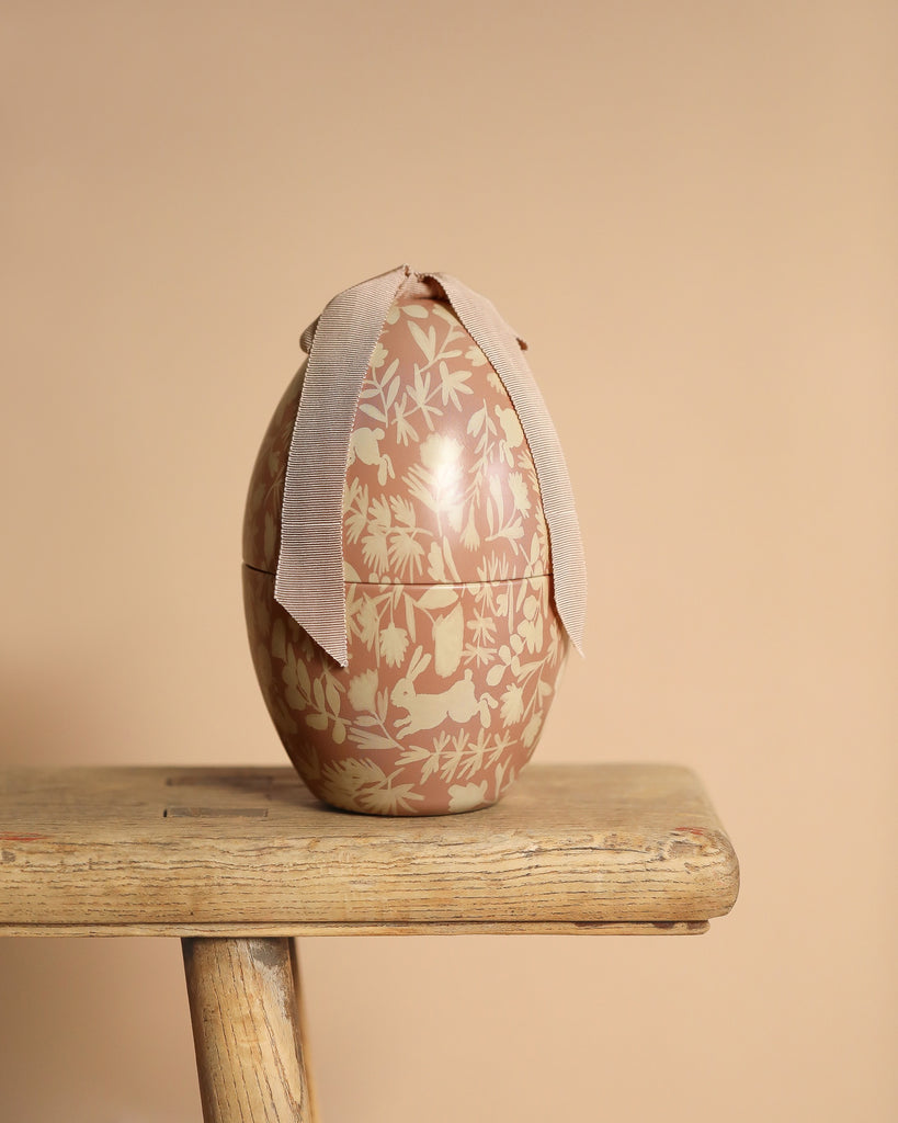 A decorative Maileg Easter Egg - Powder with a floral and leaf pattern in beige and cream tones sits elegantly on a wooden bench. Adorned with a light beige ribbon at the top, its exclusive look stands out against the soft, neutral beige background.