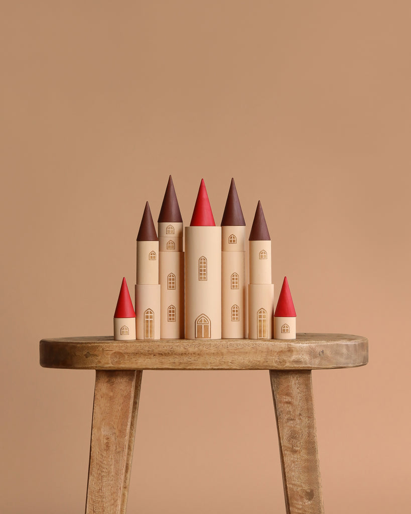 The Sabo Concept Kingdom Building Set with 7 Towers features a fairy-tale castle with red and brown roofs and arched windows, displayed on a rustic stool against a beige background—ideal for creative play with wooden blocks.