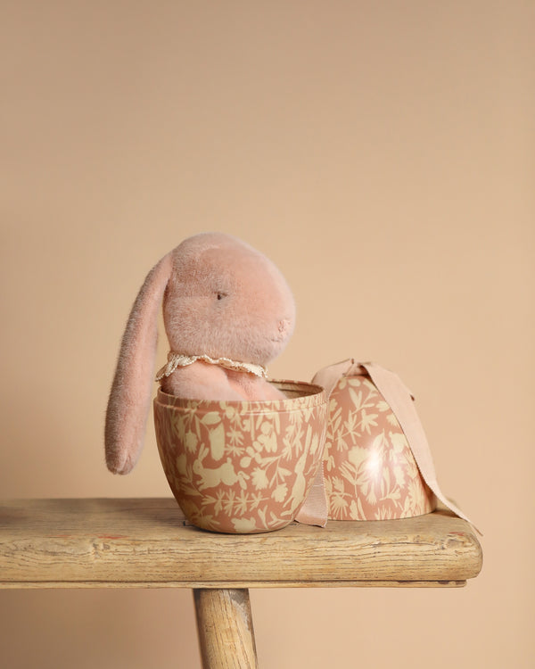 The Maileg Easter Egg With Bunny - Powder features a plush, pink bunny filled with PE pellets and floppy ears, nestled inside an open floral egg-shaped container resting on a wooden stool against a beige background.