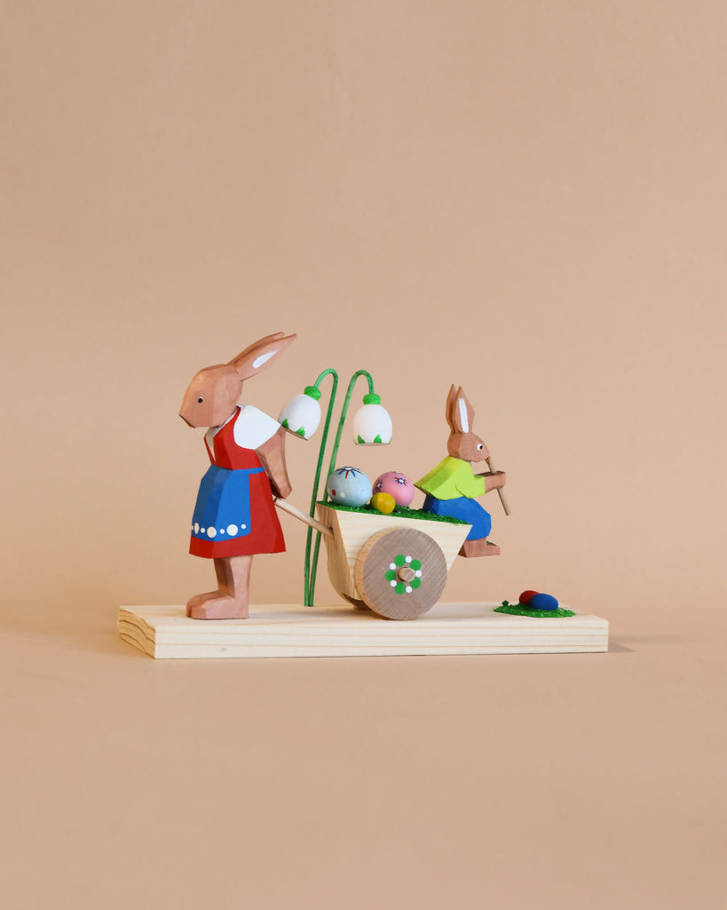 Two cherished Collectible Dregeno Easter Figures - Rabbit Mother and Son, one in a blue dress and another in green, next to a basket of colorful eggs on a wooden base against a tan background. Handcrafted in Germany.