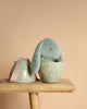 The Maileg Easter Egg With Bunny - Mint is displayed open on a wooden stool, with a plush green rabbit toy nestled inside the floral teal egg-shaped container. The lid rests beside it, all set against a soft beige backdrop.