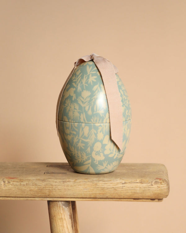 A PRE-ORDER: Maileg Easter Egg - Mint, adorned with floral patterns and a beige ribbon, rests on a rustic wooden bench against a light brown backdrop. Its exclusive appearance is highlighted by the elegant metal detailing.