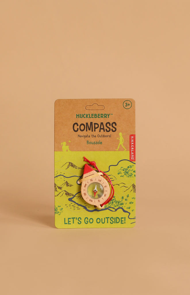 The Huckleberry Compass, made of beechwood with a red string, comes in eco-friendly packaging with green mountain art and "LET'S GO OUTSIDE!" text. Ideal for kids’ outdoor navigation, suitable for ages 3+.