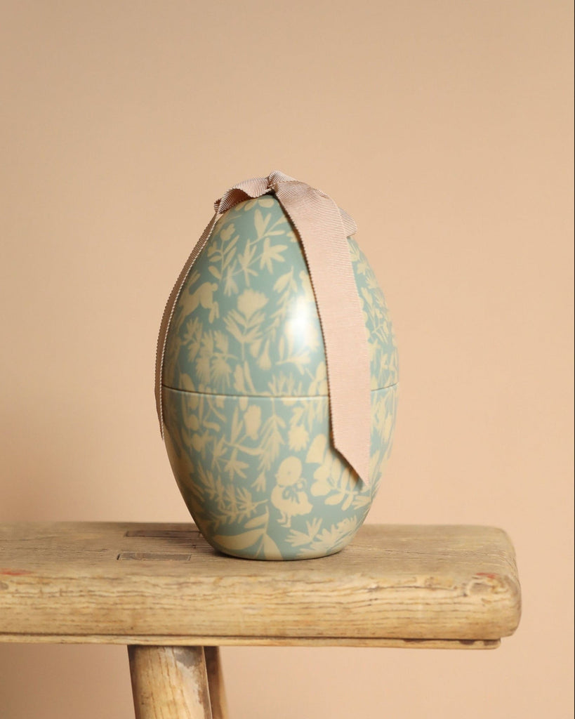 The Easter Basket Gift Set features an egg-shaped container with a light blue floral pattern and a beige ribbon bow, displayed on a wooden stool against a plain beige background.