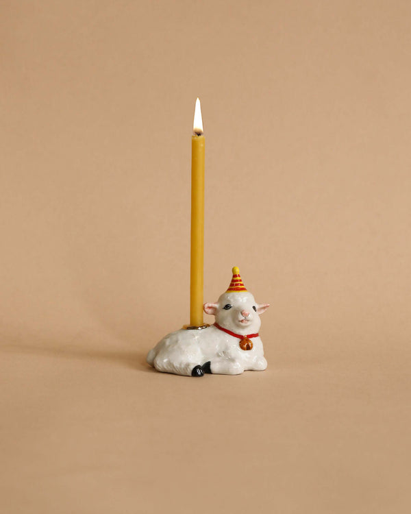 A whimsical heirloom-quality Goat Cake Topper featuring a smiling white sheep wearing a red scarf and a party hat, with a tall, lit yellow birthday candle sticking up from its back. The background is a