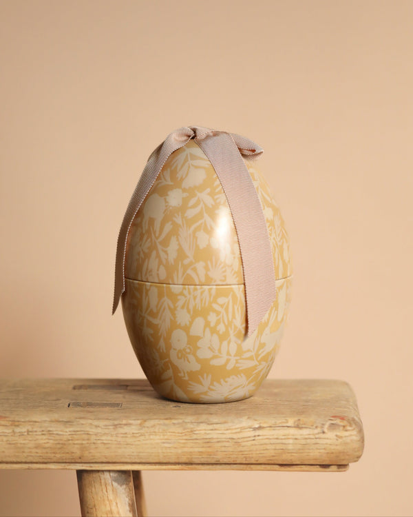 The Maileg Easter Egg - Cream Yellow, adorned with an exclusive floral pattern and beige bow, sits gracefully on a rustic wooden stool against a soft beige backdrop, providing a charming and unique look.