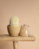 The Maileg Easter Egg With Chick features a plush yellow chick made of recycled polyester peeking from a floral-patterned egg. The open egg, with its lid adorned by a pink ribbon, rests on a wooden stool against a warm beige background.