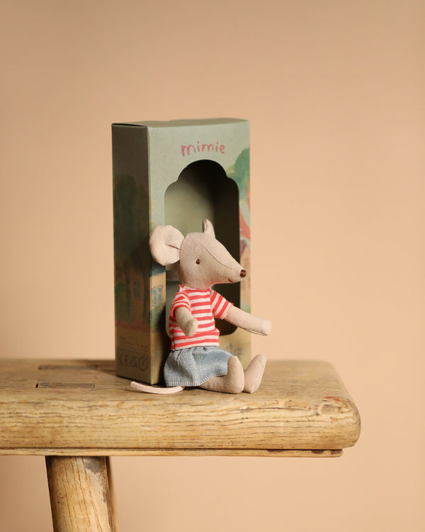 A Maileg Mouse With Striped Shirt - Big Sister, in a red striped t-shirt and blue shorts, sits on a wooden stool. Behind it is a green decoration box labeled "mimie," featuring an arched opening against a warm beige background.