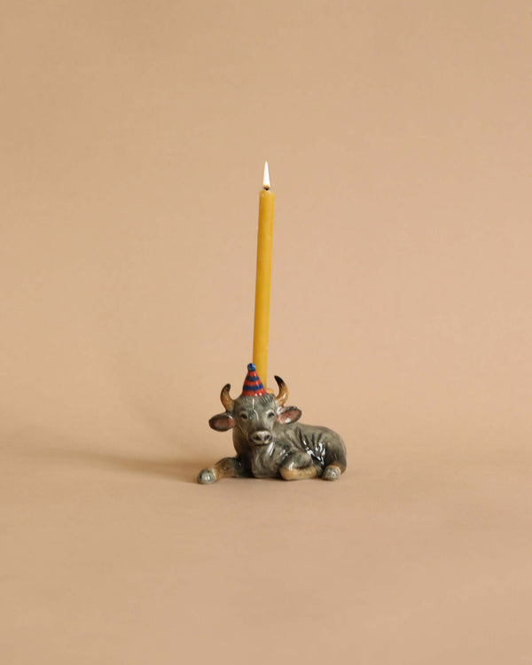 A lit yellow beeswax birthday candle supported in an Ox Cake Topper shaped like a bull, set against a plain beige background.