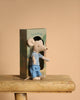 A Maileg Big Brother mouse plush in a striped shirt and blue pants stands by its "louimax" box on a wooden stool, beige background. Ideal as a companion for your Maileg Big Brother and Sister Afternoon Tea Set.
