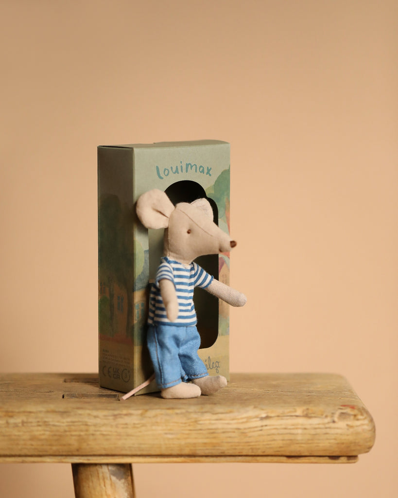 The Maileg LouisMax Mouse - Big Brother, in a striped shirt and blue pants, sits on a wooden stool with a decorative "LouiMax" box featuring whimsical illustrations behind it against a calming beige background.