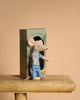 A small Maileg mouse toy dressed in blue pants and a striped shirt stands in front of the Maileg LouiMax Bathroom Set box, which is placed on a wooden stool against a plain beige background.