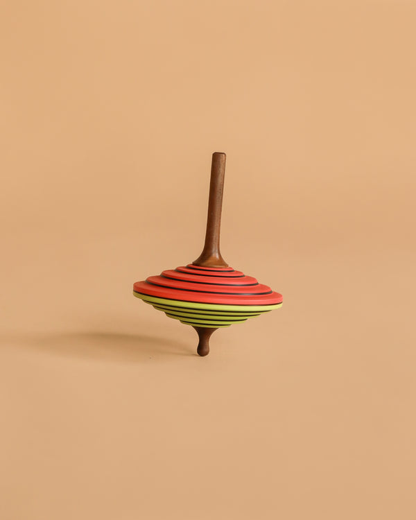 The Mader Tukan Spinning Top in green and red features vibrant, layered rings and stands upright on a beige background—a classic wooden toy inspired by Austrian craftsmanship.