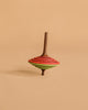 The Mader Tukan Spinning Top in green and red features vibrant, layered rings and stands upright on a beige background—a classic wooden toy inspired by Austrian craftsmanship.